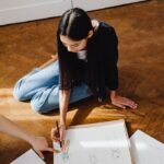 A young woman sits on the floor sketching fashion designs with pencil and paper.